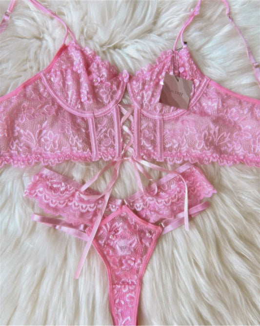 Vintage lingerie set with pink lace bralette and thong, floral embroidery, and ribbon details on a soft fabric background.